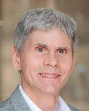 Headshot of a person with short gray hair, smiling, wearing a light-colored shirt and blazer.