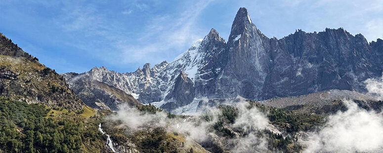 Jagged rocky mountain peaks rising above green forested slopes, with low clouds drifting through the valley below.