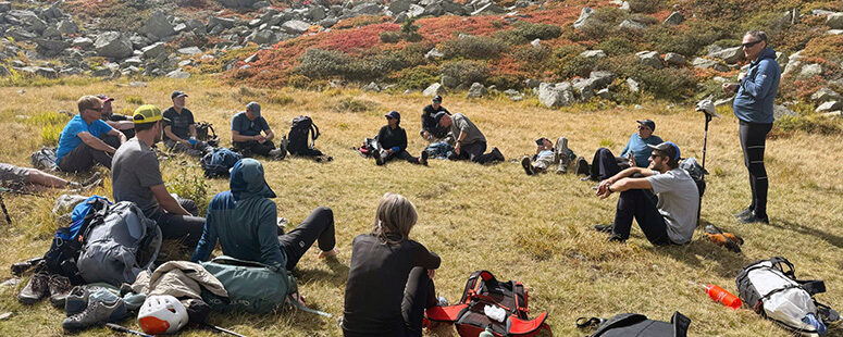 A group of hikers sitting in a circle on a grassy alpine meadow, surrounded by backpacks, rocks, and autumn-colored vegetation.