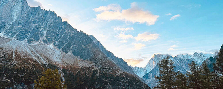Snow-dusted mountains and rocky cliffs at sunset, with warm light on the peaks and trees in the foreground.